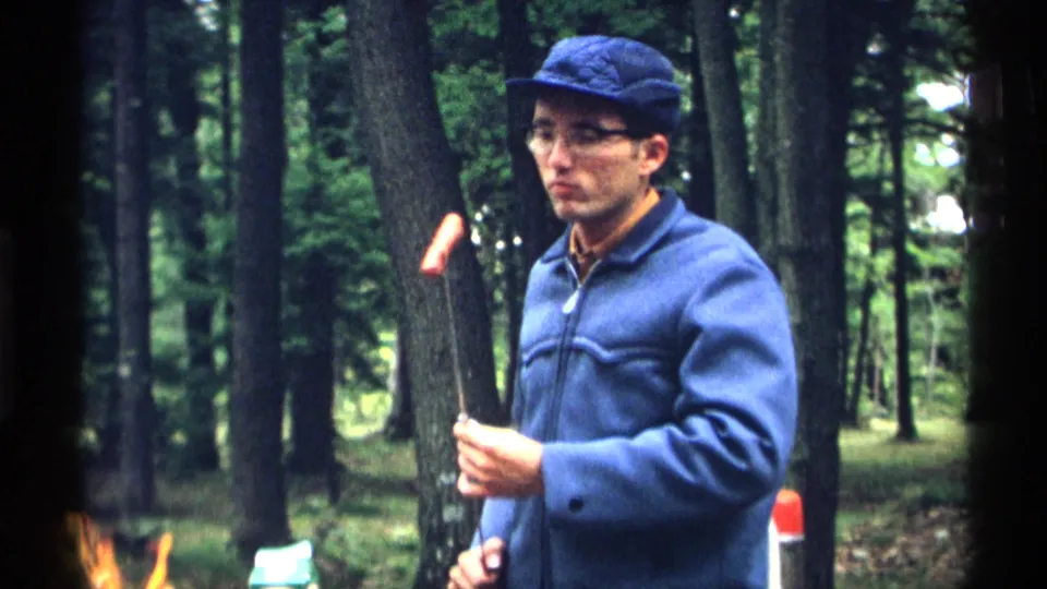 In 1971 Wisconsin 8mm footage, a man in a blue cap and jacket holds a roasting stick with a hot dog near trees at a campsite.