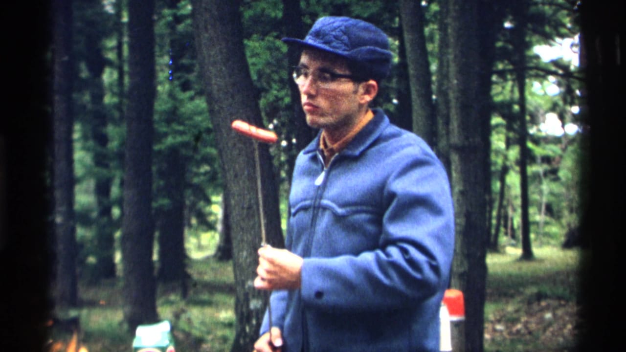 Man in blue jacket holds a hot dog on a roasting stick near trees.