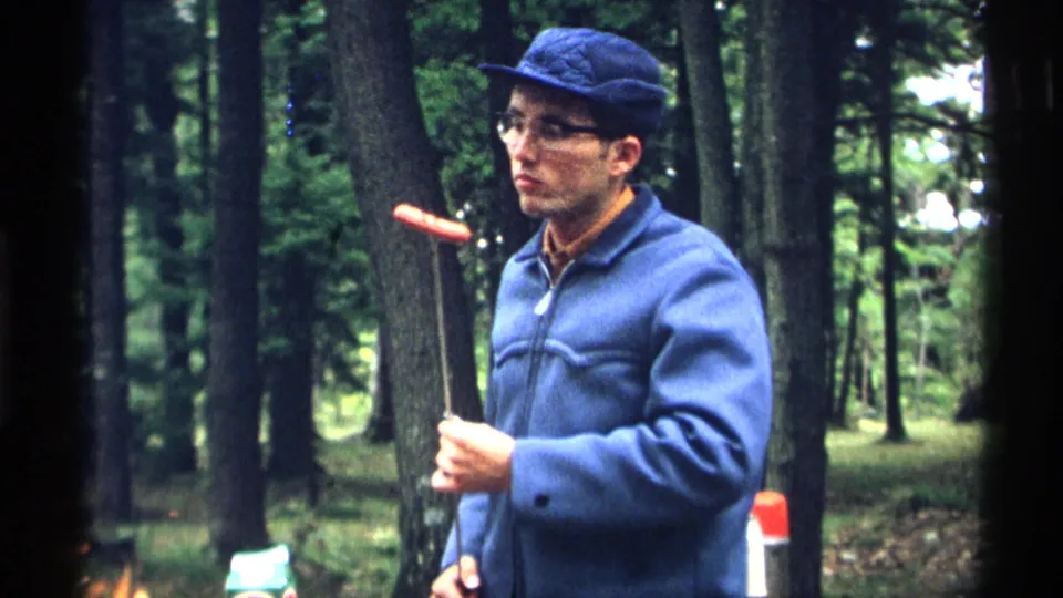 The man in a blue jacket and cap watches a hot dog on a roasting stick in the wooded Wisconsin campsite footage.