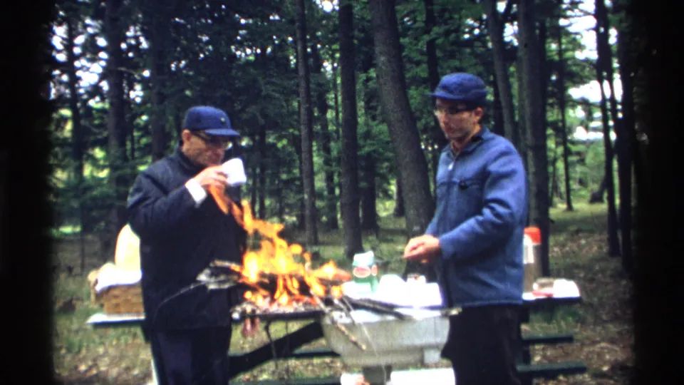 Two adult men are beside a campfire in 1971 Wisconsin, one drinking from a white mug while the other tends a roasting stick.