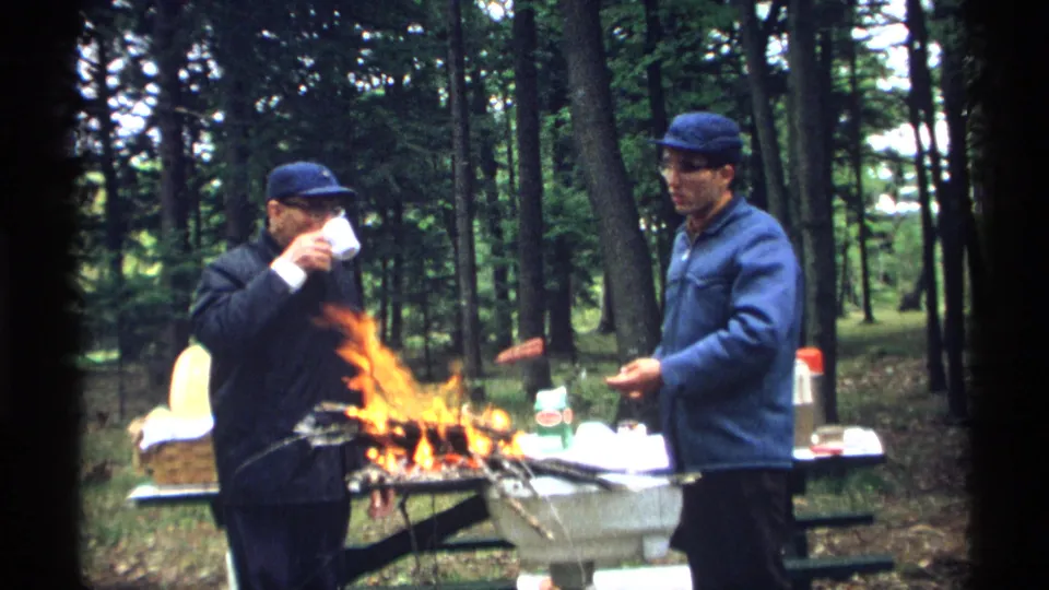 At the wooded campsite, one man drinks from a mug as another man in a blue jacket holds a hot dog near the campfire.