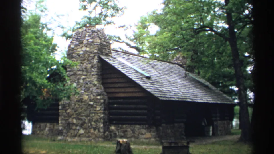 1971 Wisconsin frame of the Brunet Island park log structure, with chimney, roofline, trees, and grass visible.