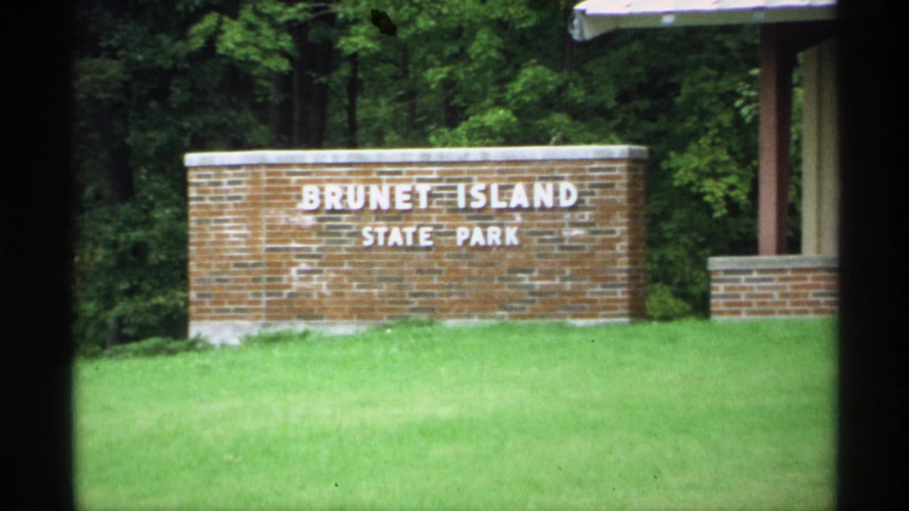1971 Wisconsin 8mm frame showing the Brunet Island State Park brick entrance sign in front of lawn and dense trees.
