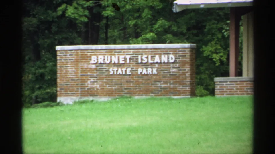 1971 Wisconsin 8mm frame showing the Brunet Island State Park brick entrance sign in front of lawn and dense trees.
