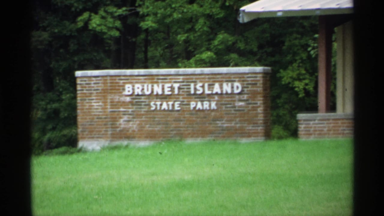 1971 Wisconsin home-movie view of the Brunet Island State Park sign with green grass, wooded background, and nearby shelter structure.