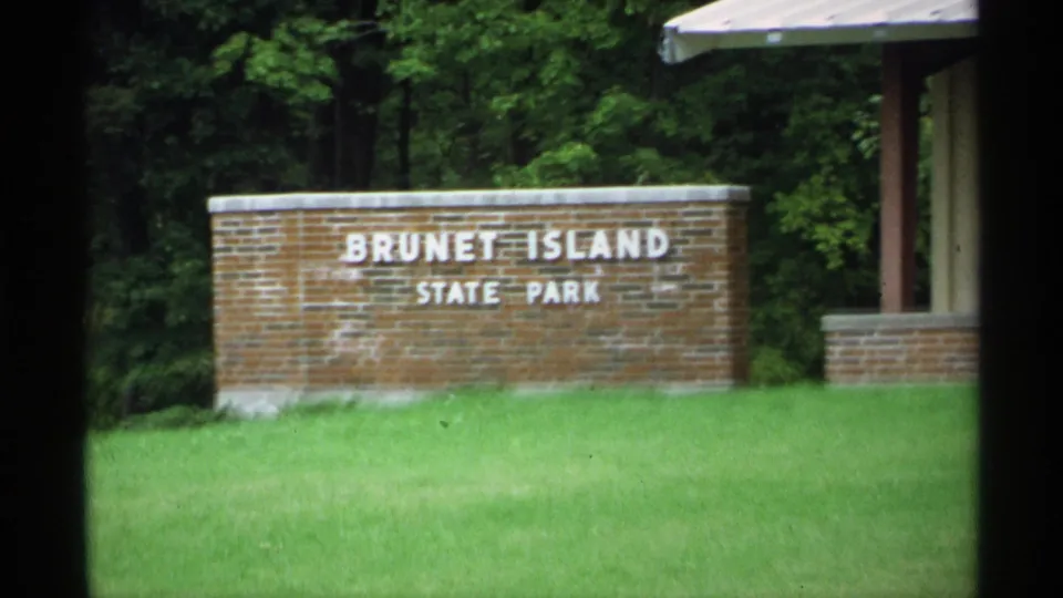 1971 Wisconsin home-movie view of the Brunet Island State Park sign with green grass, wooded background, and nearby shelter structure.