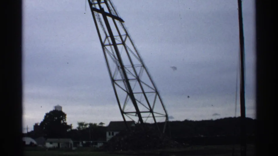 1971 Wisconsin 8mm frame showing a tall industrial crane, overcast sky, distant low buildings, and a water tower.