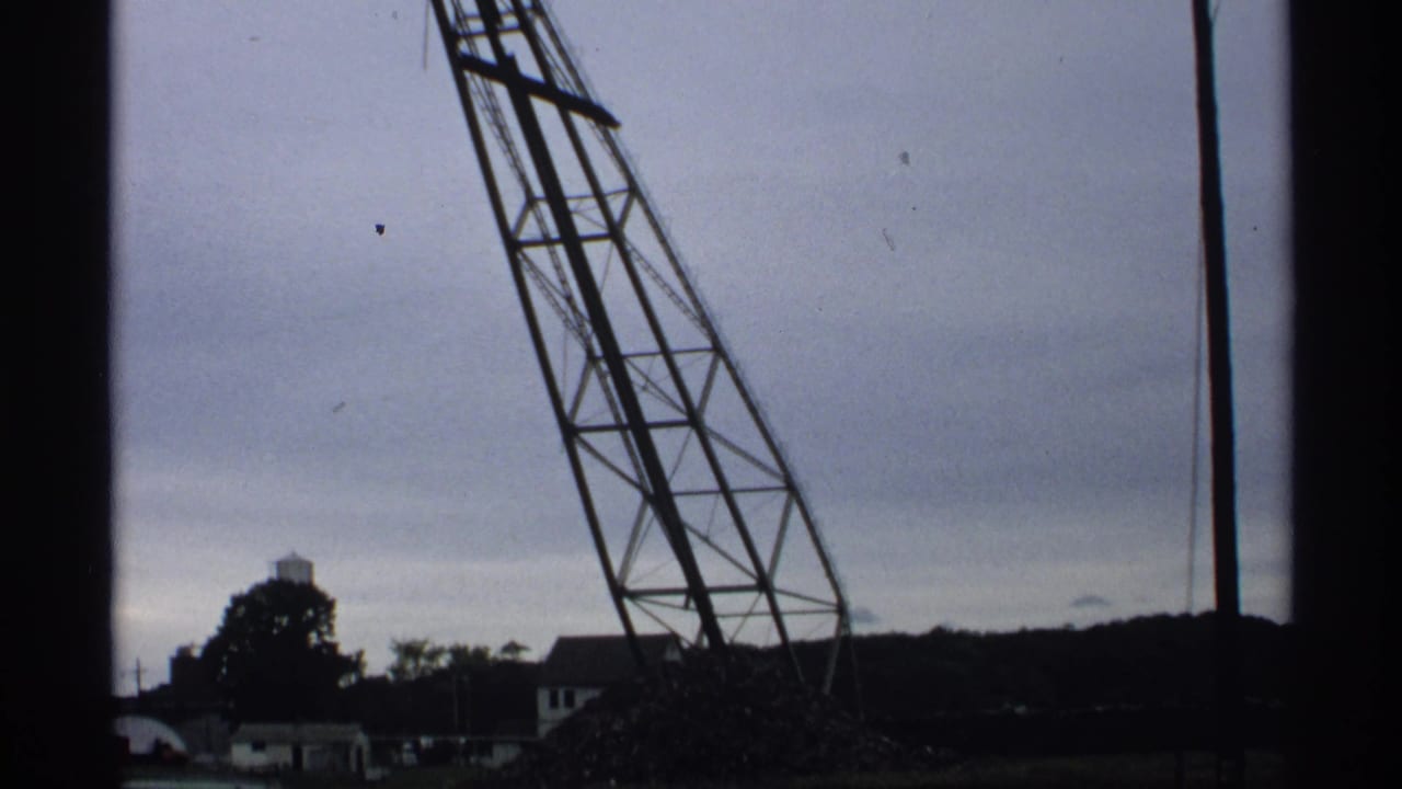 1971 Wisconsin 8mm frame showing a damaged lattice gantry leaning near distant buildings and a water tower under overcast sky.