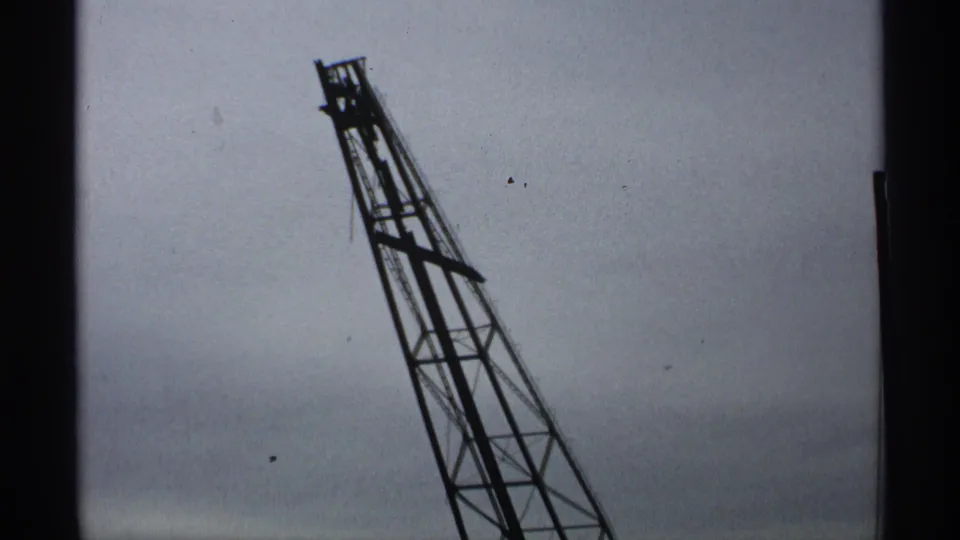 Final 8mm frame of the damaged gantry leaning across a muted cloudy sky, with dust specks and soft exposure.