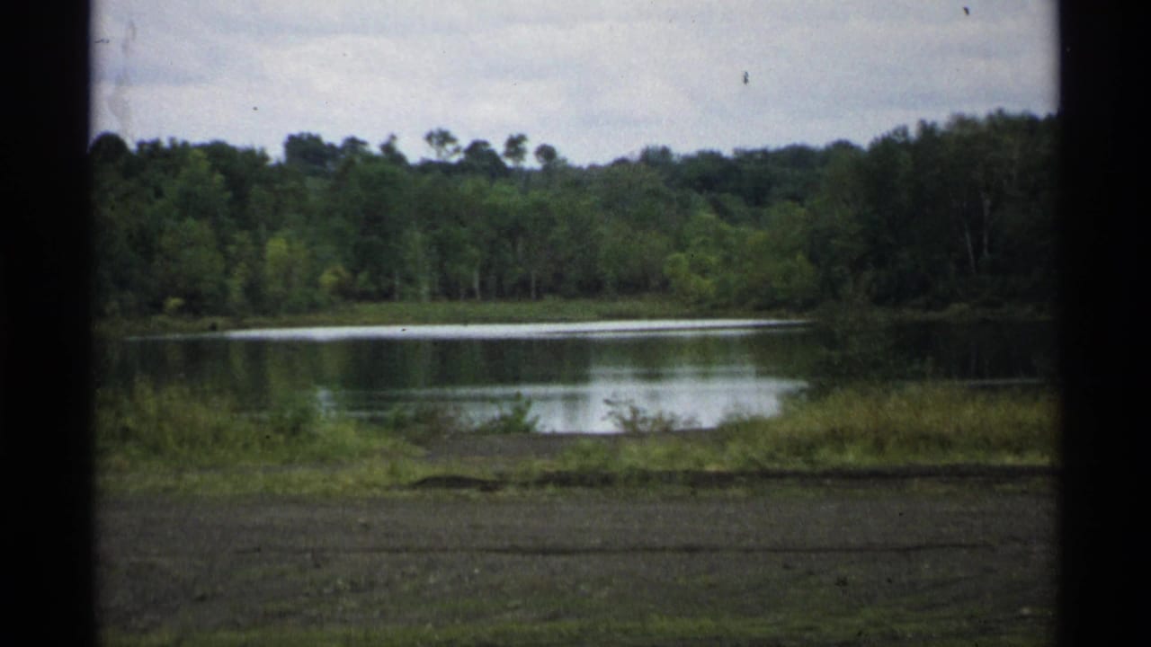 1971 Wisconsin 8mm frame showing a calm lake, wooded shoreline, grassy bank, and cloudy sky from a roadside home-movie view.