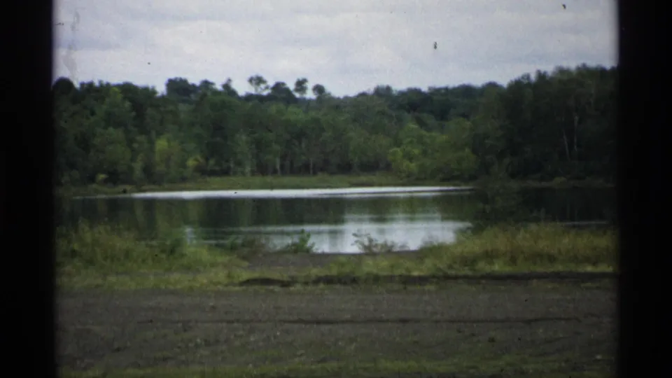 1971 Wisconsin 8mm frame showing a calm lake, wooded shoreline, grassy bank, and cloudy sky from a roadside home-movie view.