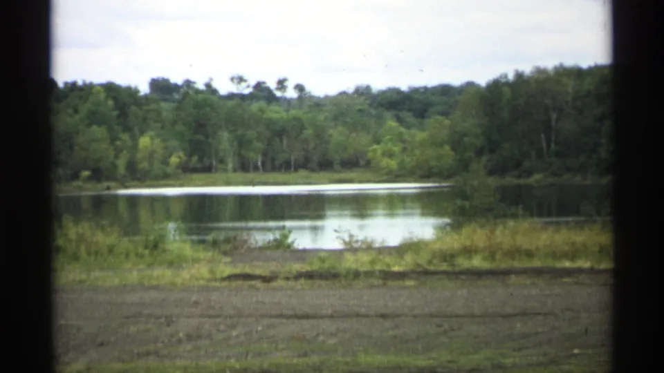 8mm Wisconsin travel frame showing lake water, dense trees, low grass, and an overcast sky in a quiet rural landscape.