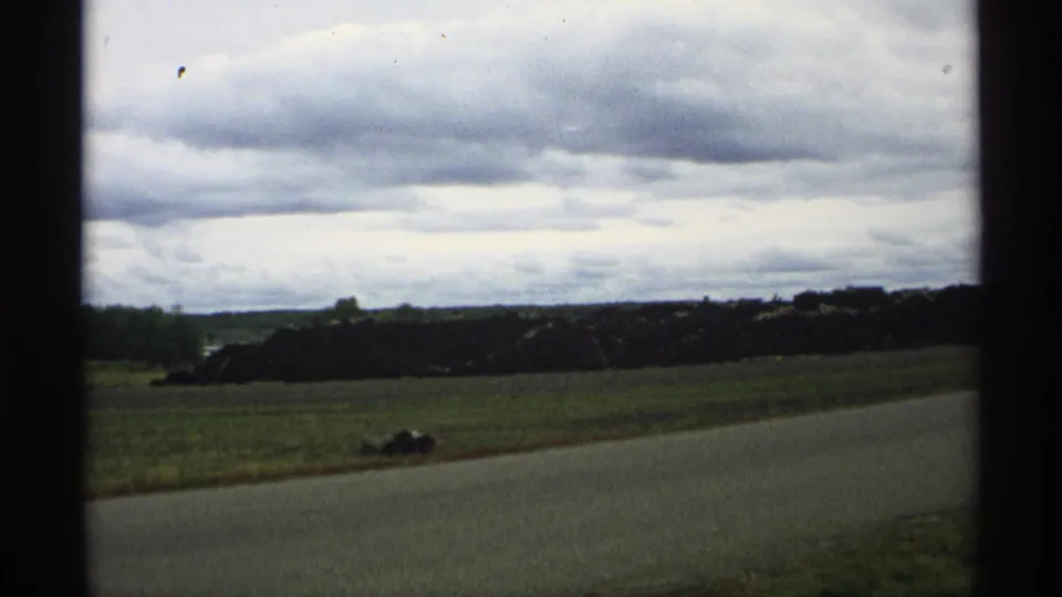 1971 Wisconsin roadside view with an open field, large cut-log pile, low horizon, and heavy overcast sky.
