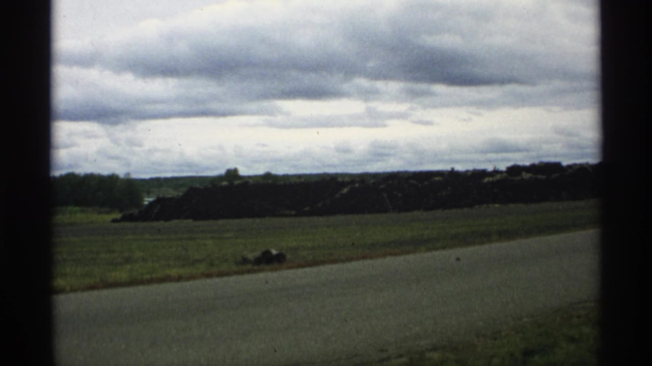 1971 Wisconsin 8mm frame showing a rural roadside, open grass field, and dark gravel or stone pile beneath a cloudy sky.