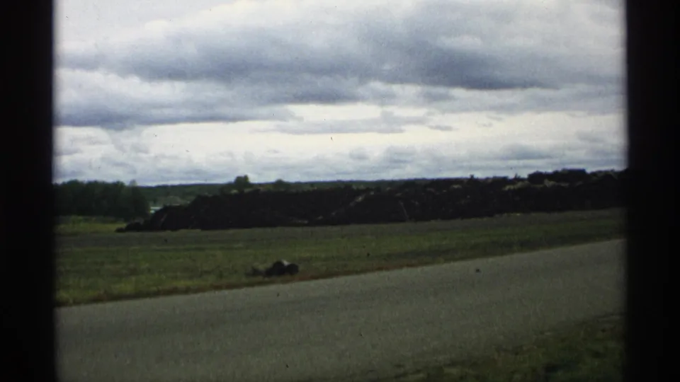1971 Wisconsin 8mm frame showing a rural roadside, open grass field, and dark gravel or stone pile beneath a cloudy sky.