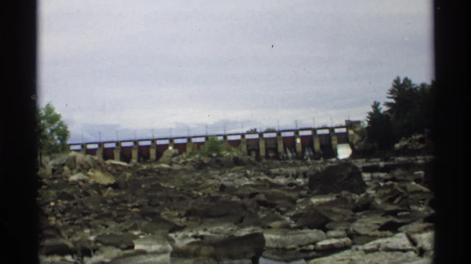 1971 Wisconsin 8mm frame of a low dam crossing a rocky riverbed, with water gates and trees along the shoreline.