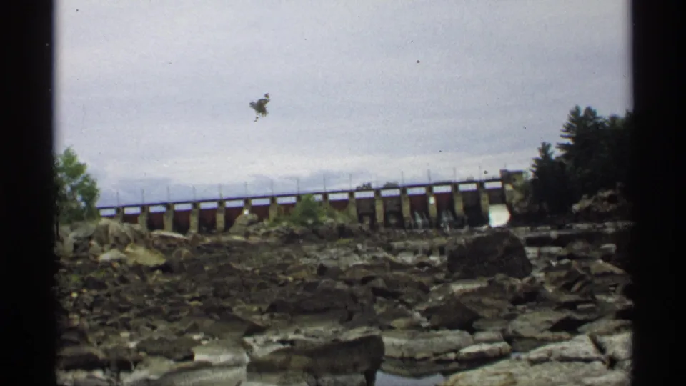 A 1971 Wisconsin home-movie view of a dam over a rocky riverbed, with trees at the right bank and muted overcast light.