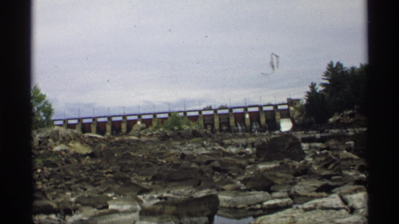 Low dam or bridge crossing a rocky riverbed under an overcast sky.
