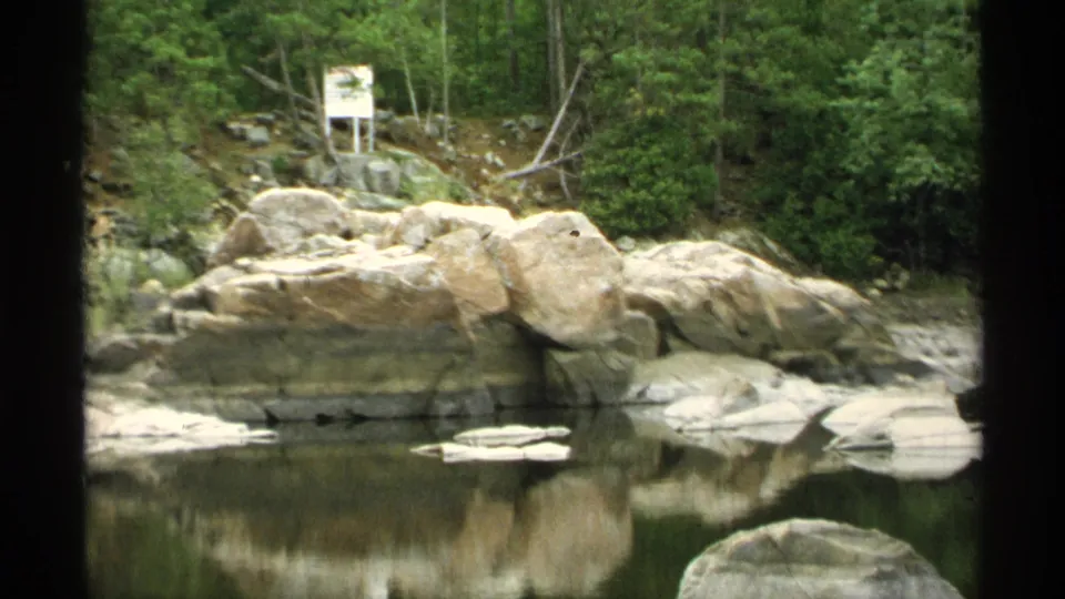 1971 Wisconsin 8mm view of large shoreline stones and forest reflected across a quiet pond or river pool.