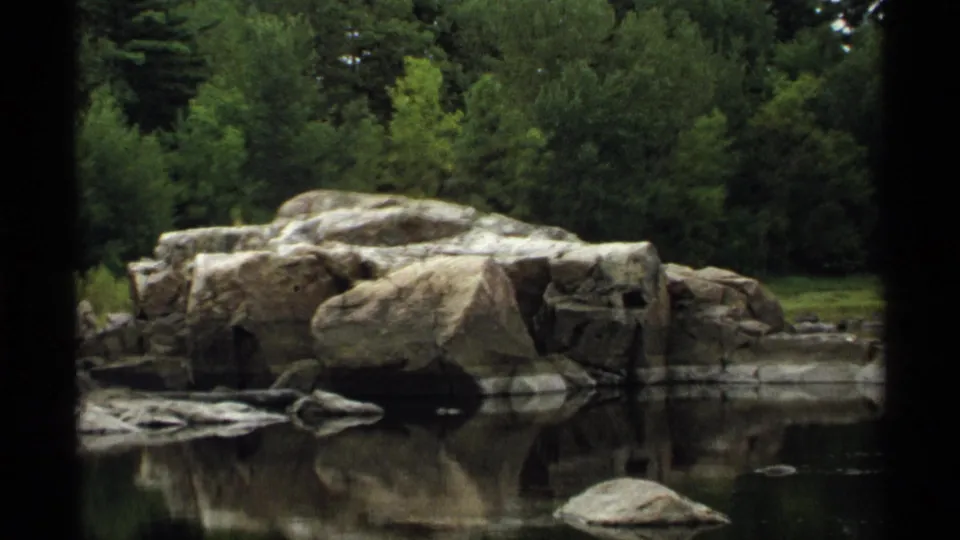 1971 Wisconsin home-movie frame showing a broad rock formation mirrored in still water, backed by dense green woods.