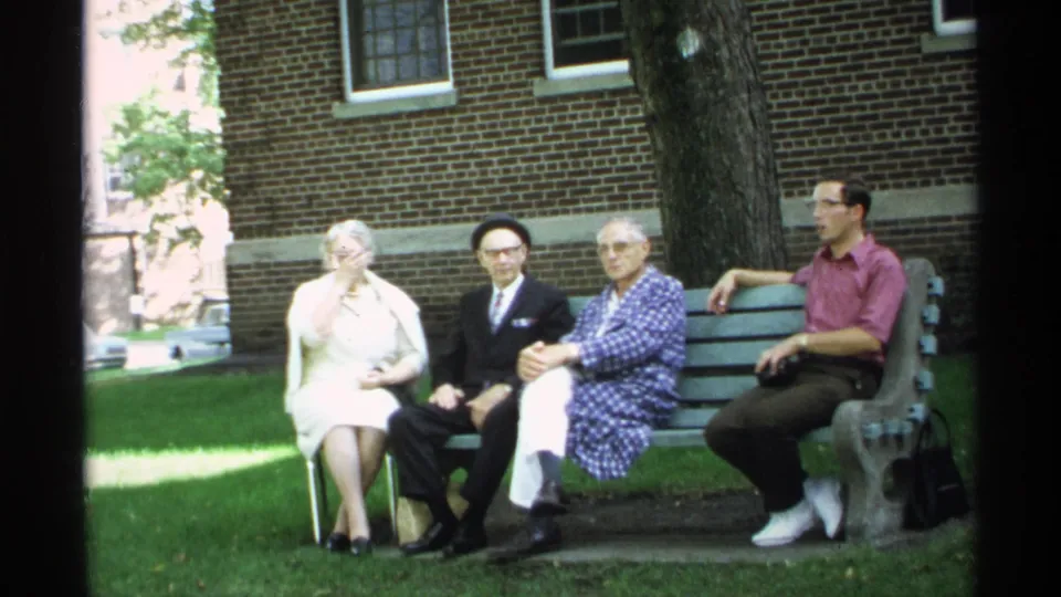 Four adults pose on a green park bench in Wisconsin, with grass, a large tree, and a brick building behind them.