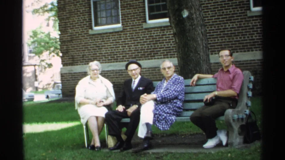 A posed 1971 Wisconsin home-movie frame shows four adults seated on a green bench outside a brick building.