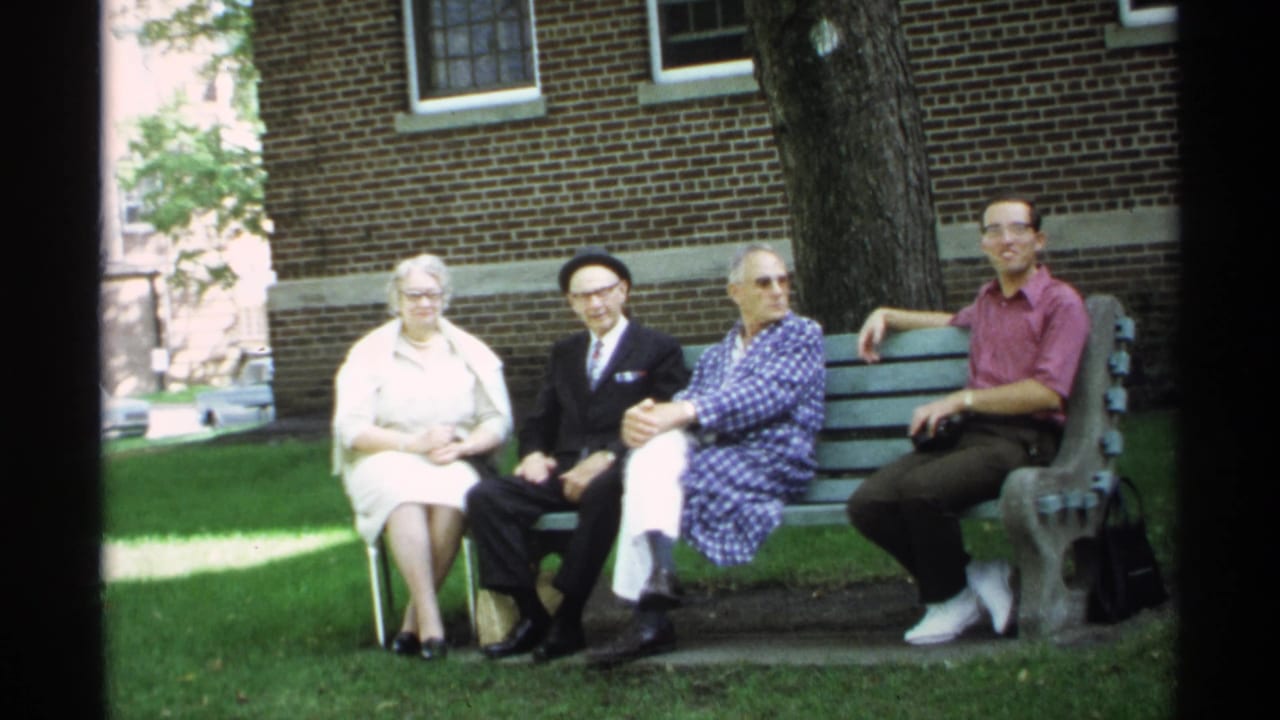 1971 Wisconsin 8mm frame showing four adults seated on a park bench beneath trees near a brick building.