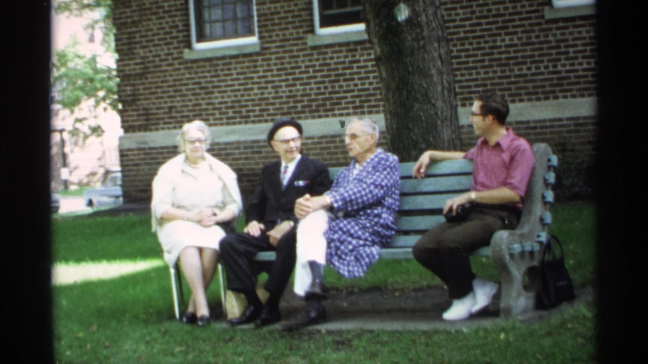 Four adults remain seated on a bench, with trees and brick wall behind them.