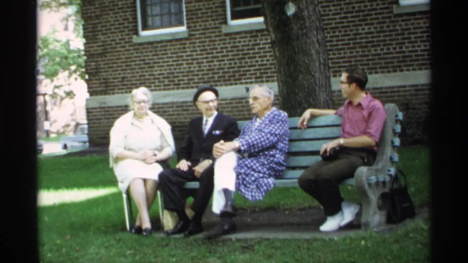 Four family members sit on a green bench in a Wisconsin park setting, framed by grass, trees, and a brick building.
