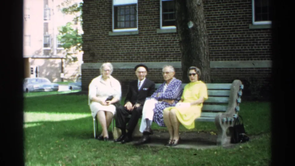 1971 Wisconsin home-movie frame with three older adults and a woman in yellow seated together on a park bench.