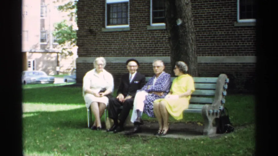 Four adults sit together on a green bench in a 1971 Wisconsin park scene, with a brick building and trees behind them.