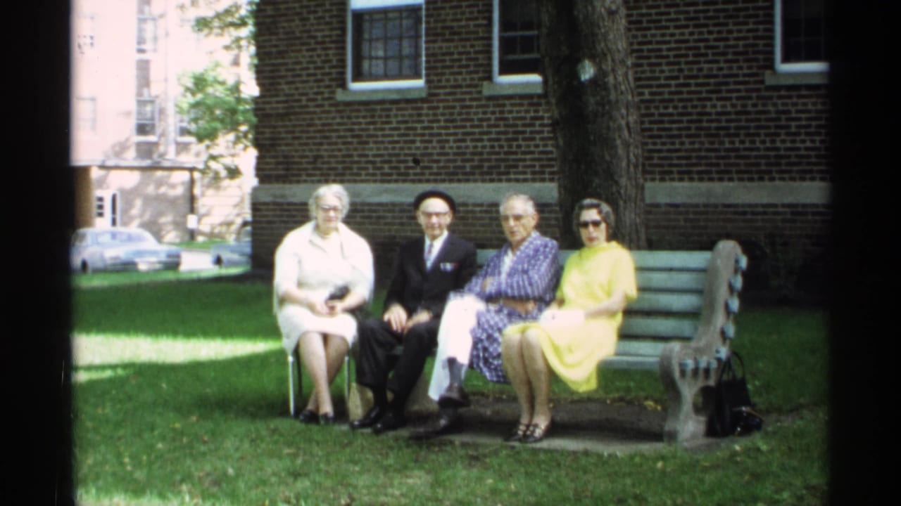 In 1971 Wisconsin home-movie footage, four older adults sit posed on a bench with grass, a tree, brick wall, and parked car behind them.