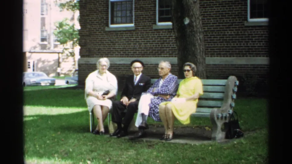 The 8mm frame shows four older adults seated on a bench in Wisconsin, including a woman in yellow and a man in a dark suit facing the camera.