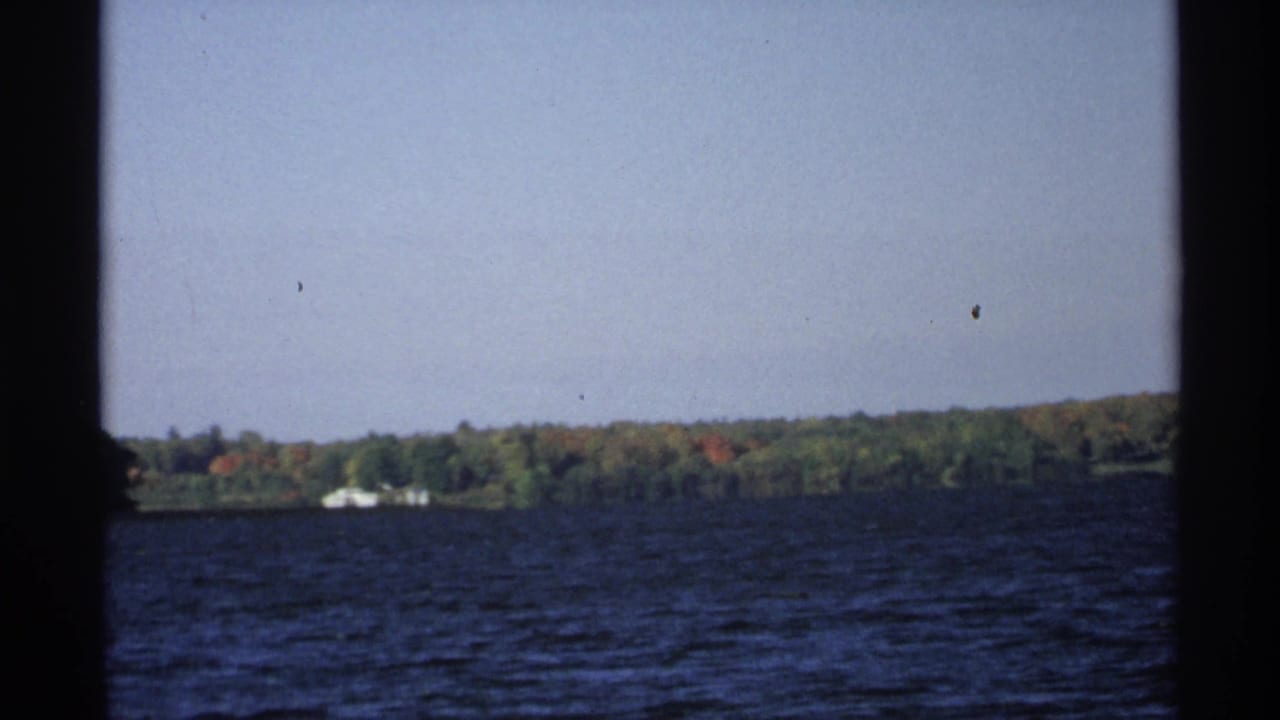 Open lake and distant wooded shore framed by dark vertical edges, with small light structures near the trees.