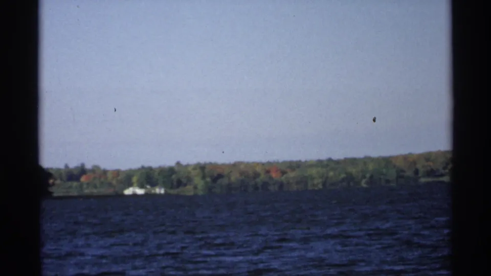 1971 Wisconsin lake view with rippled water, a tree-lined shore, and small light shoreline structures visible through dark frame edges.
