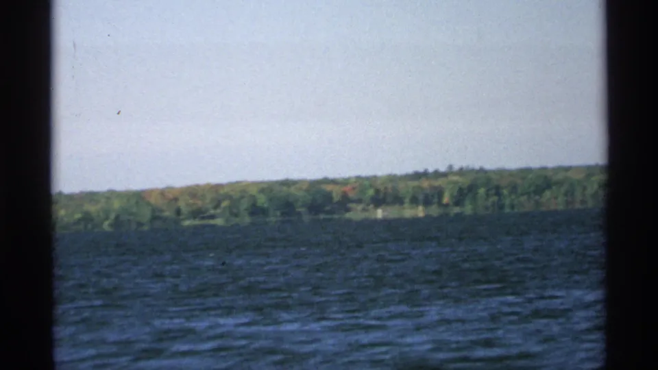 1971 Wisconsin home-movie frame showing rippled blue water and a distant green shoreline under a pale sky.