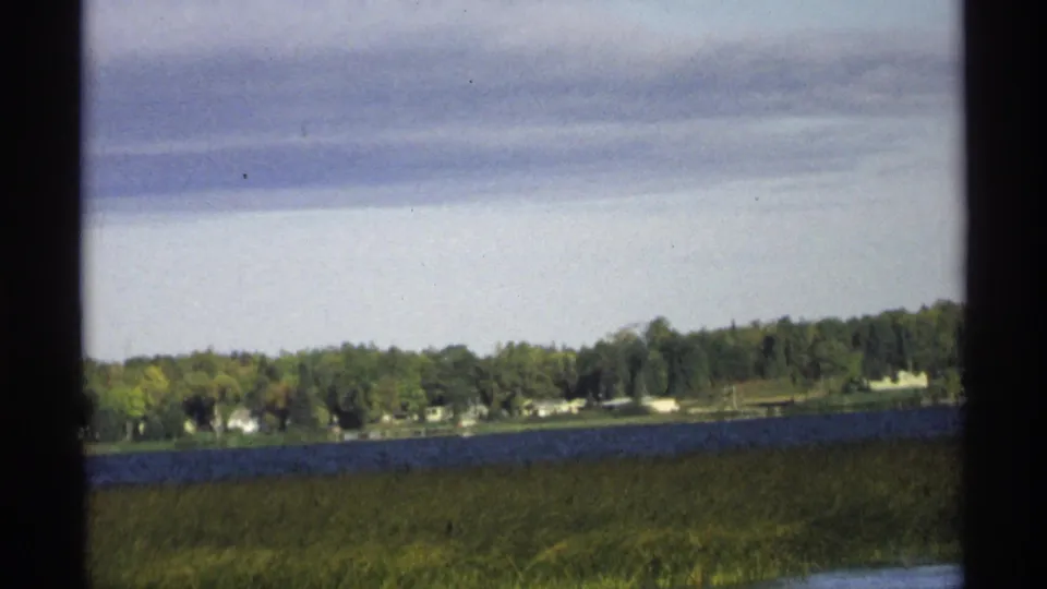 1971 Wisconsin lake view with marshy grass in front, blue water, wooded shore, and small distant houses among trees.