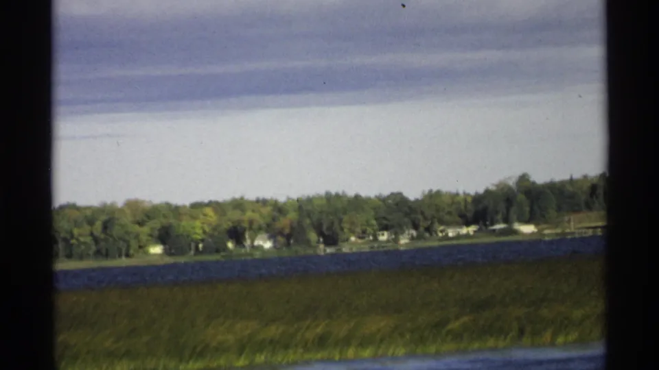 Slightly tilted 1971 Wisconsin 8mm frame of a lake edge, grassy foreground, wooded shoreline, and small distant buildings.