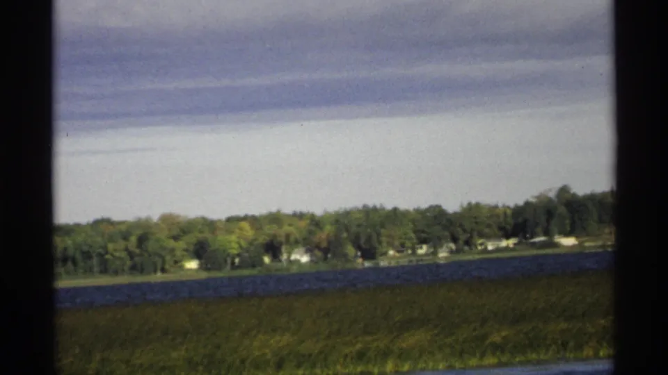 1971 Wisconsin lake scene with marsh grass, rippled blue water, a wooded far shore, and cloudy sky in a soft 8mm frame.