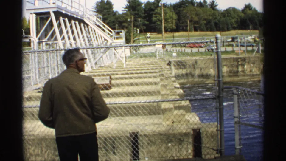 1971 Wisconsin home-movie frame showing an adult man by a fence observing Pokegama Dam's concrete spillway and water.