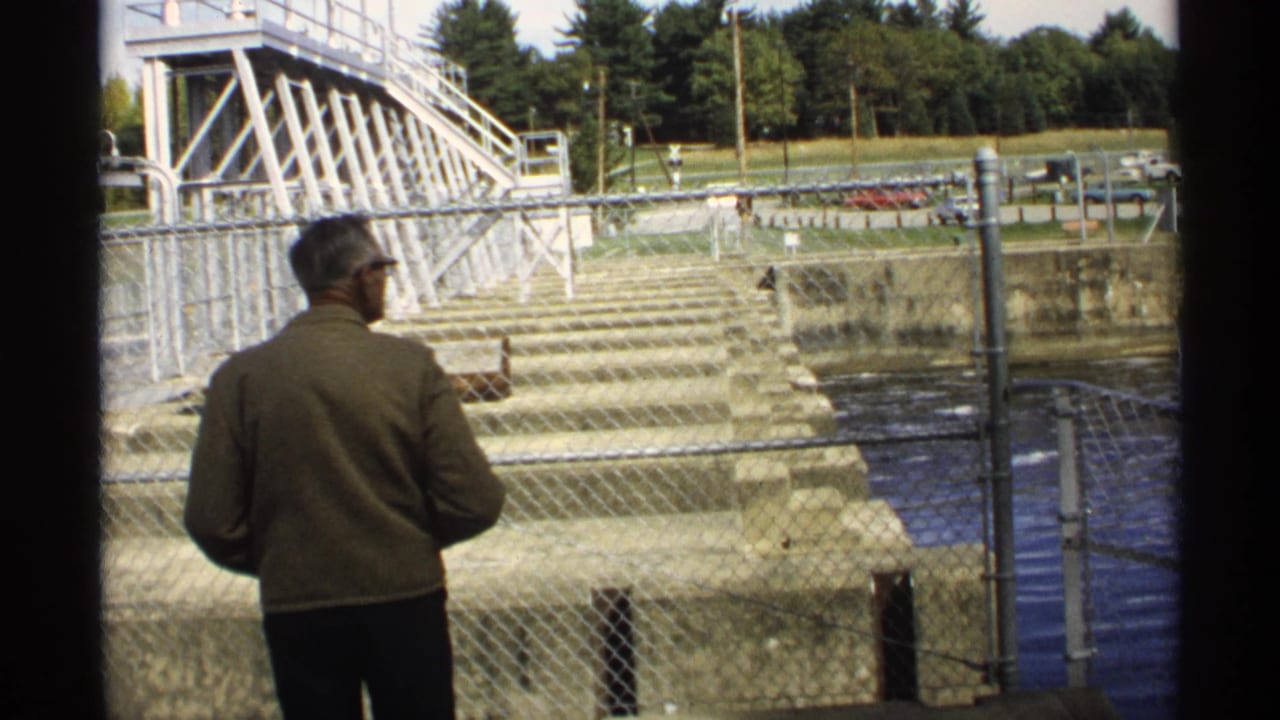 Man in a brown jacket stands by a chain-link fence near a concrete dam.