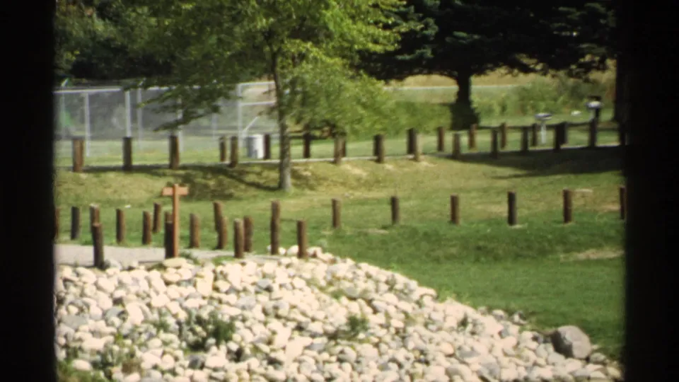 The camera shows a grassy Wisconsin park area with wooden posts, a small cross, rocks, fencing, and trees on 8mm film.