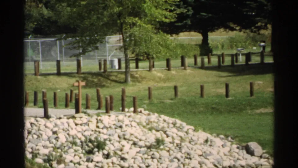 A 1971 Wisconsin park view shows rocks in the foreground, a small cross, wooden posts, fencing, trees, and open grass.
