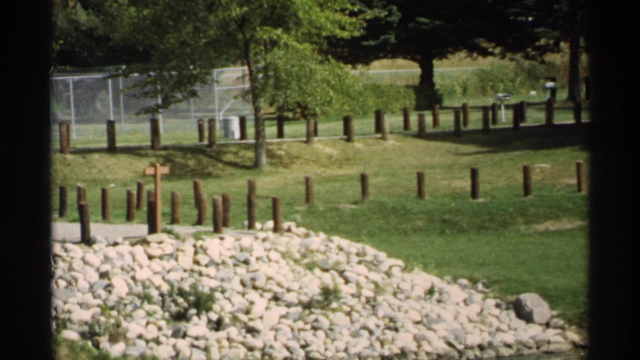 Park lawn with rocks, wooden posts, trees, and a small cross near a path.