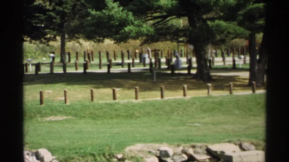 1971 Wisconsin park view with mature trees, rows of wooden posts, a paved path, and small distant visitors in the background.