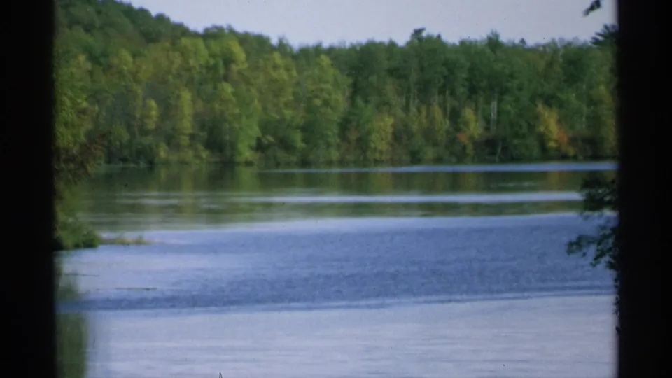 1971 Wisconsin home-movie view of calm lake water and a dense green tree line, partially framed by dark foreground edges.