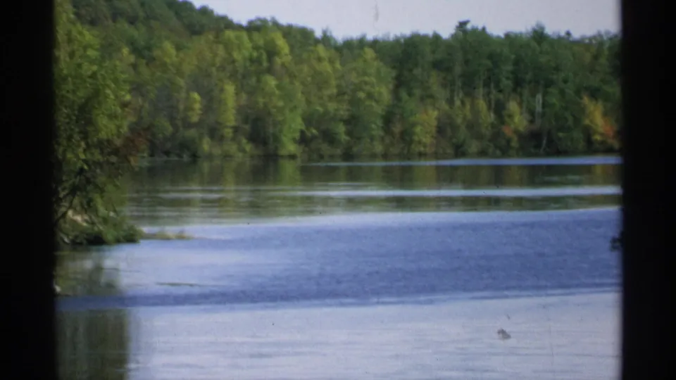 1971 Wisconsin lakeside frame showing calm water, a wooded far shore, and a small visible object or post near the lower right waterline.