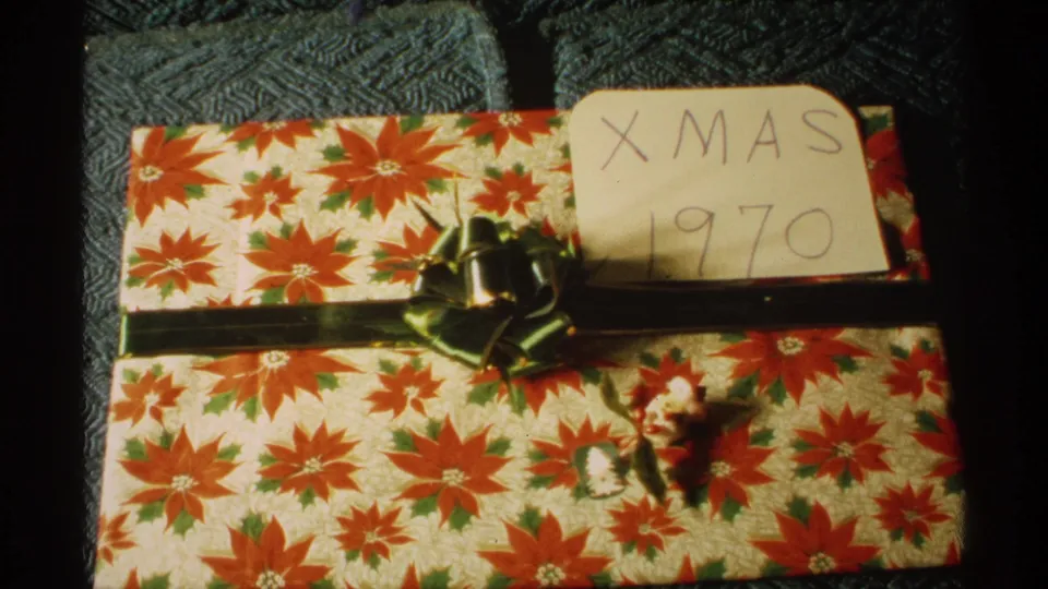 Home-movie frame showing a wrapped Christmas gift on textured upholstery, with green ribbon and a handwritten card reading Xmas 1970.