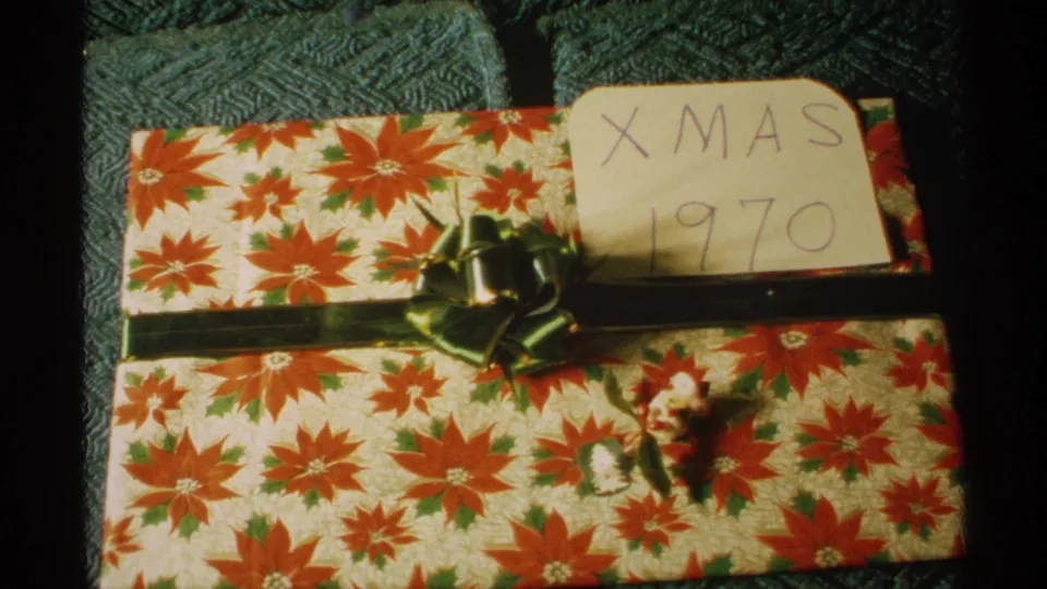 Vintage home-movie frame of a holiday package with red floral wrapping, green ribbon, bow, and a card marked Xmas 1970.