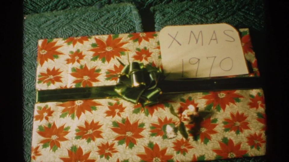 Home-movie frame showing a wrapped Christmas gift on textured upholstery, with red floral paper, green ribbon, and a partly visible Xmas 1970 card.